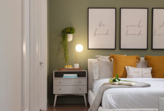 Earthy room decor featuring a natural sage green wall, greenery, wood flooring, yellow throw pillow, and white bed linens.