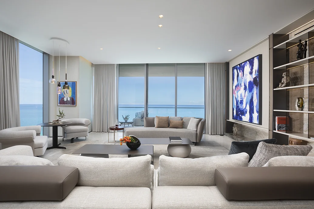 Luxury oceanfront living room with cream seating, built-in shelving, colorful artwork, floor-to-ceiling windows, and Miami water views