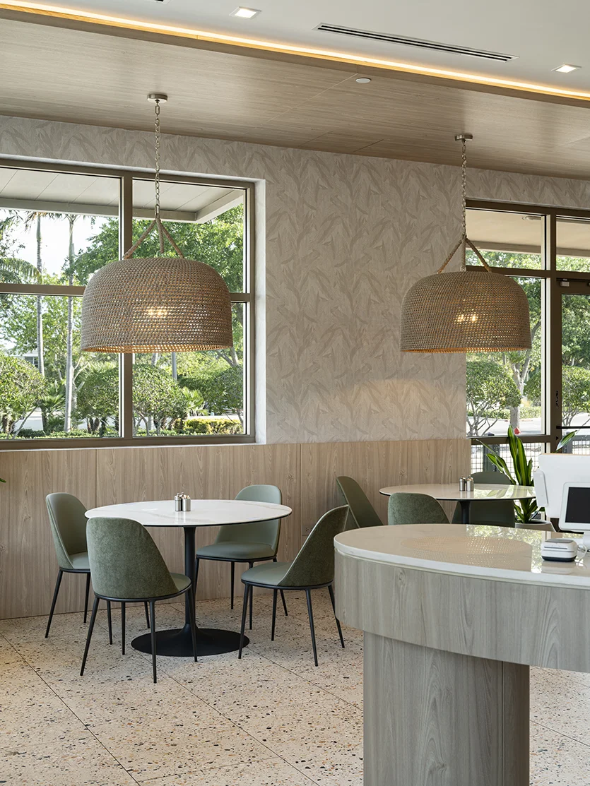 Bright restaurant dining area with round tables, sage green chairs, woven pendant lights, a curved service counter, and large windows