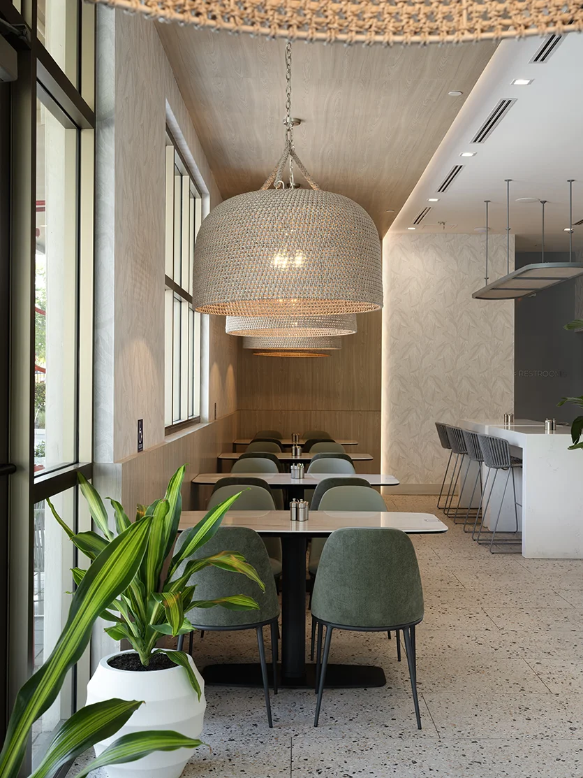 Modern Fort Lauderdale restaurant dining room with sage green chairs, woven pendant lights, large windows, and a white bar counter