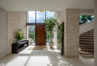Modern foyer entryway in a Coral Gables whole home renovation featuring travertine stone walls, floor-to-ceiling glass windows, a rich wood front door, and a curved staircase.