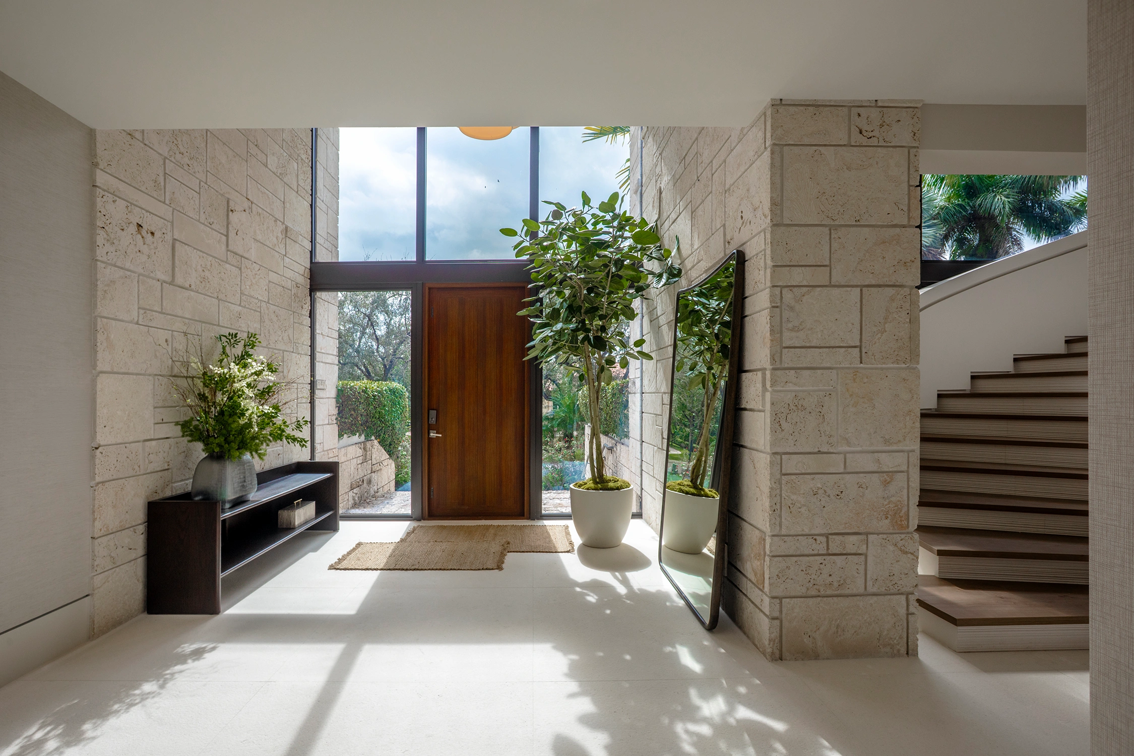 Modern foyer entryway in a Coral Gables whole home renovation featuring travertine stone walls, floor-to-ceiling glass windows, a rich wood front door, and a curved staircase.