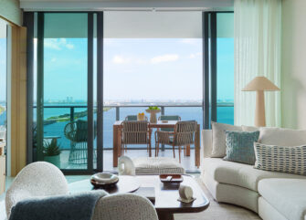 Luxury bayfront living room at One Paraiso Miami featuring a curved neutral sofa, organic decor, and floor-to-ceiling glass sliding doors opening to a balcony with panoramic ocean views.