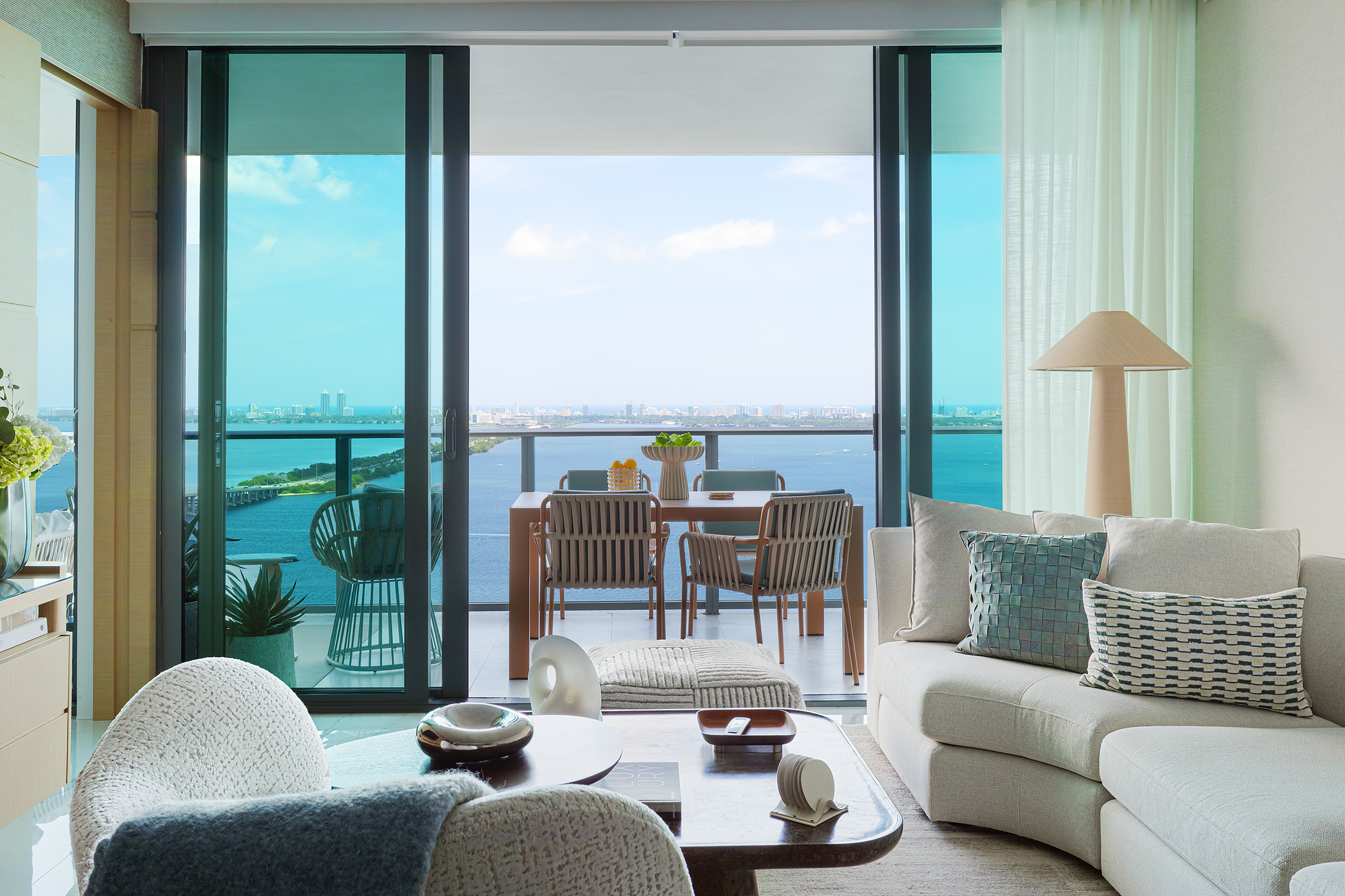 Luxury bayfront living room at One Paraiso Miami featuring a curved neutral sofa, organic decor, and floor-to-ceiling glass sliding doors opening to a balcony with panoramic ocean views.