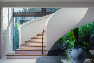 Modern curved staircase after renovation featuring smooth white balustrades, warm wood treads, and natural travertine wall detailing.