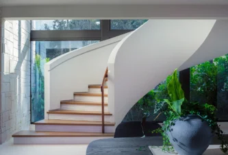 Modern curved staircase after renovation featuring smooth white balustrades, warm wood treads, and natural travertine wall detailing.